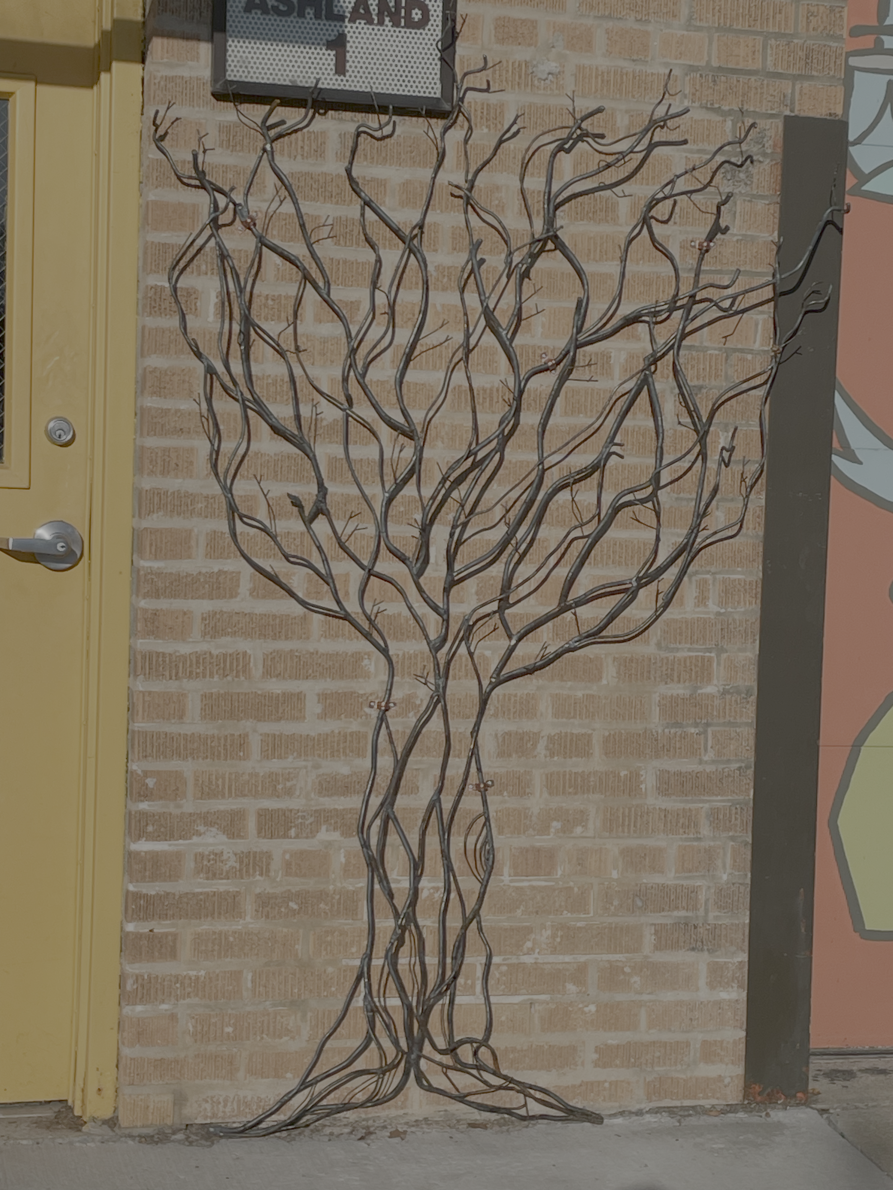 Metal sculpture of tree trunk and branches against brick wall between door and mural on front of Rex's Place