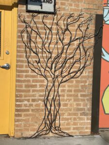 Metal sculpture of tree trunk and branches on brick wall between door and mural