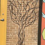 Metal sculpture of tree trunk and branches on brick wall between door and mural
